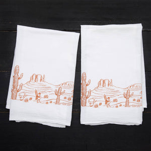 two rectangular dish towel with an orange illustration of a desert landscape. The design features cactuses and mountains.