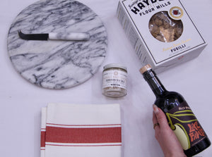Hand holding a bottle of olive oil, box of pasta, hand towel, sea salt, marble cheese table