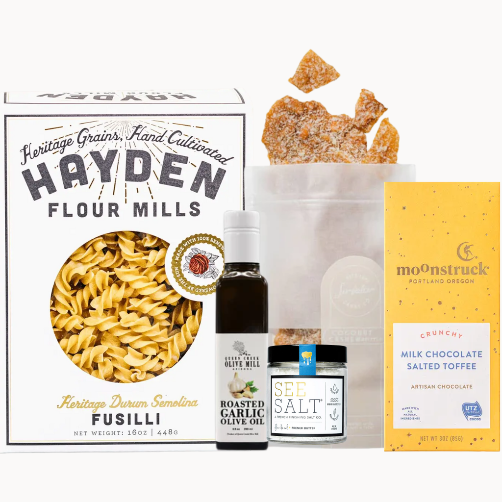 Assorted food products including a box of Hayden Flour Mills fusilli pasta, a bottle of olive oil, a jar of salt, and a box of Moonstruck chocolate on a white background.
