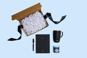 black pen, black notebook, black tumbler, candy jar, open box filled with crinkle paper