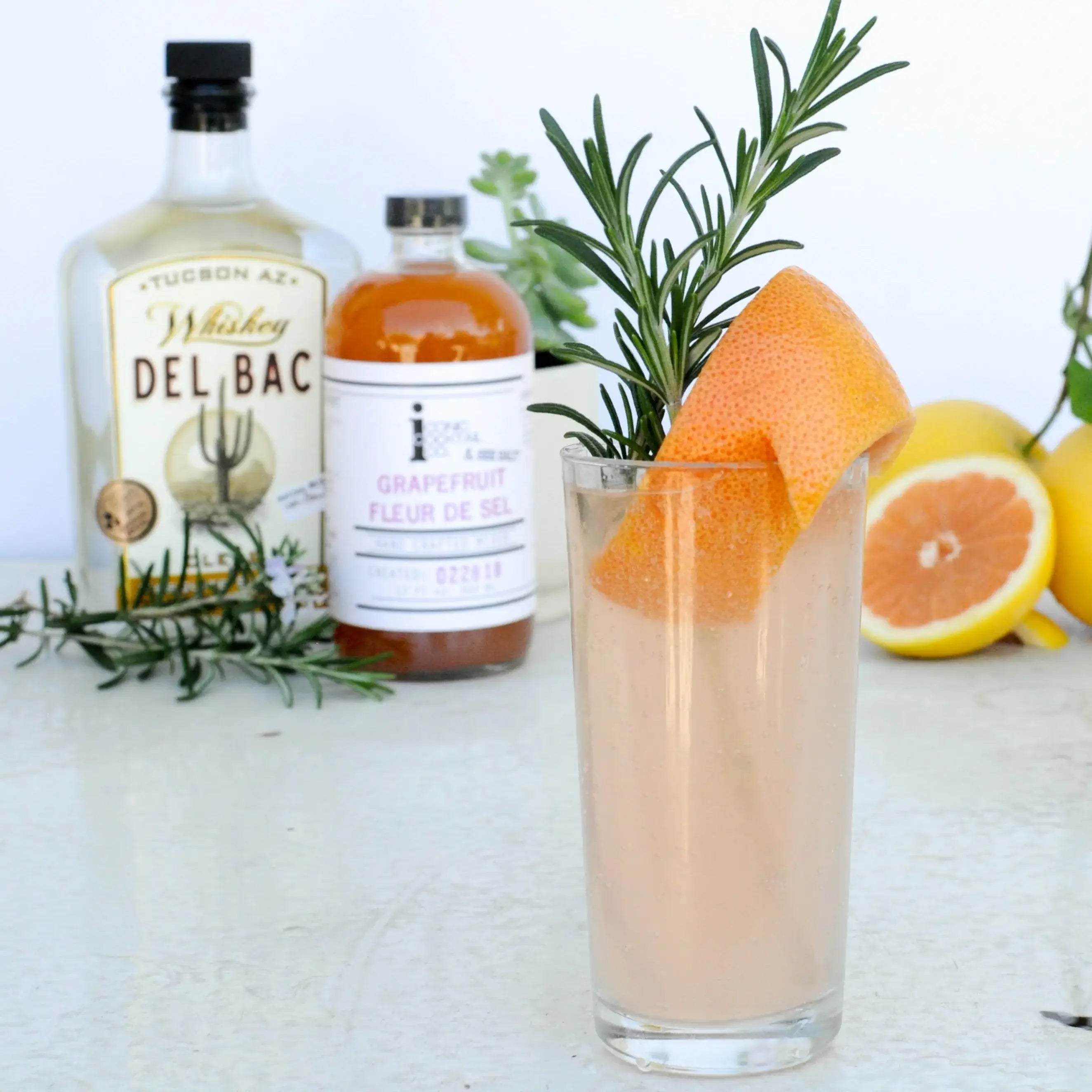 Glass of pink cocktail with grapefruit and rosemary, surrounded by bottles and citrus fruits on a white background