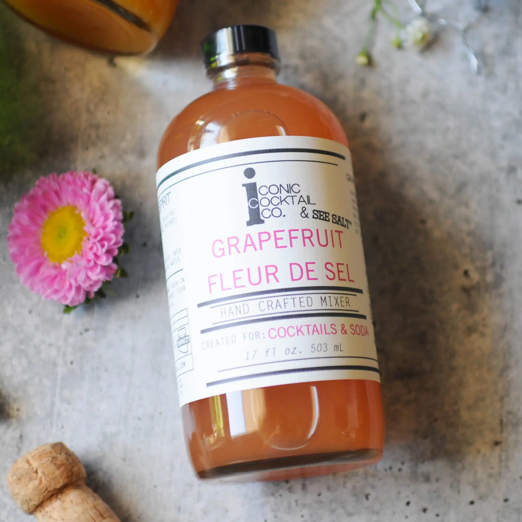 Bottle of Grapefruit Fleur de Sel mixer from Iconic Cocktail Co. on a textured surface with a pink flower.