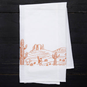 A rectangular dish towel with an orange illustration of a desert landscape. The design features cactuses and mountains.