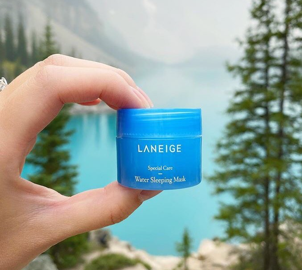 A hand holding a small blue jar, with a natural background. . The jar has the Laneige logo and text "Water Sleeping Mask" written on the front in white lettering.