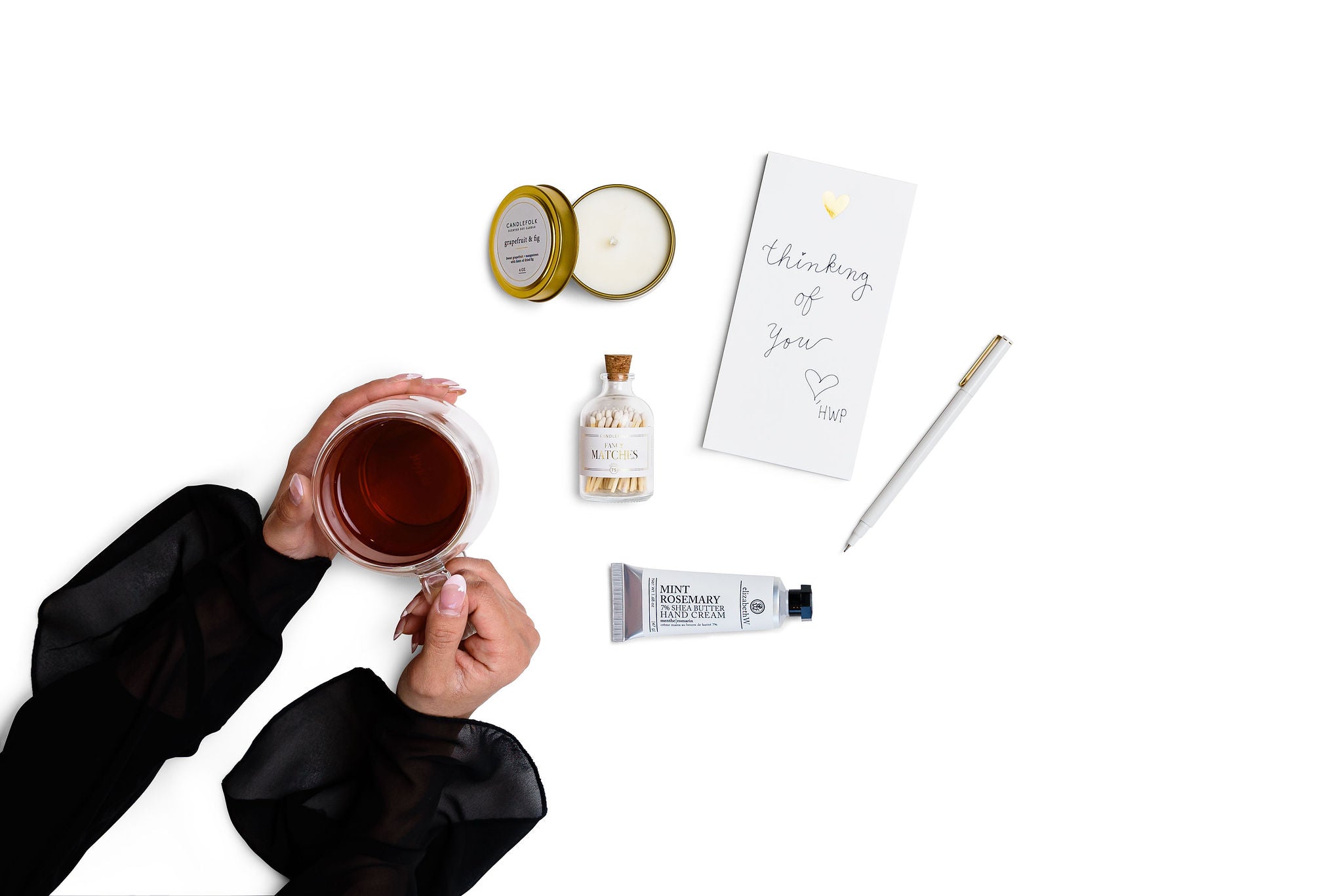 hands holding a glass cup fith tea, open candle with golden lid on top, matches jar, hands cream, white open pen, notepad with thinking oh you wirtten on it