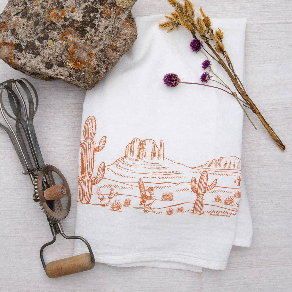 A rectangular dish towel with an orange illustration of a desert landscape. The design features cactuses and mountains., a mineral  rock, a dessert flower and a vintage manual mixer.